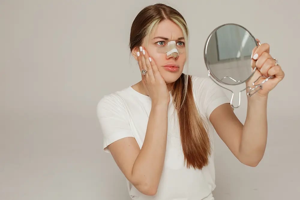 woman recovering from rhinoplasty surgery a young woman with a surgical bandage and splint on her nose looks into a handheld mirror